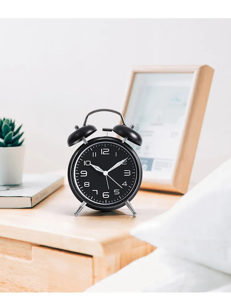 Large black metal alarm clock with luminous hands and face, 4 inches, on a wooden bedside table