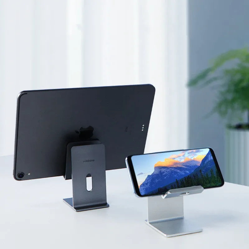 Dark grey alloy mobile phone and tablet holder with adjustable clamp on a wooden desk.