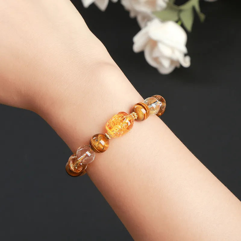 A close-up of a beaded bracelet with yellow banded Tiger's Eye and translucent citrine 10mm beads and a metallic Pi Xiu charm.