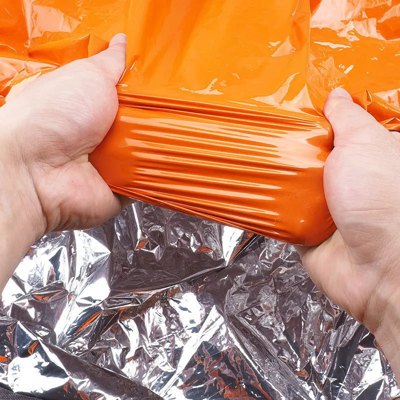 Orange emergency survival blanket unfolded showing reflective inner surface, with folded sleeping bag and tent beside it in nylon bag