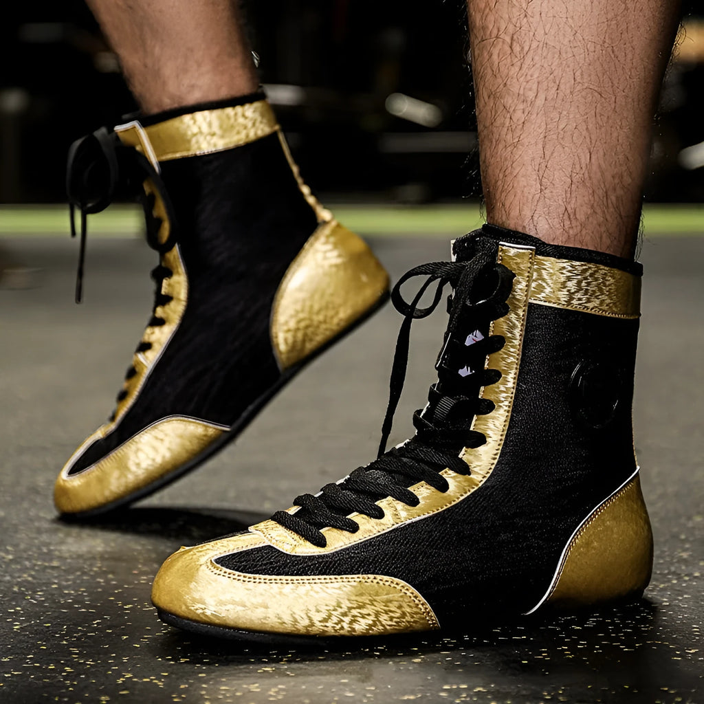 Pair of black and gold professional boxing shoes with mesh panels on a gym floor.