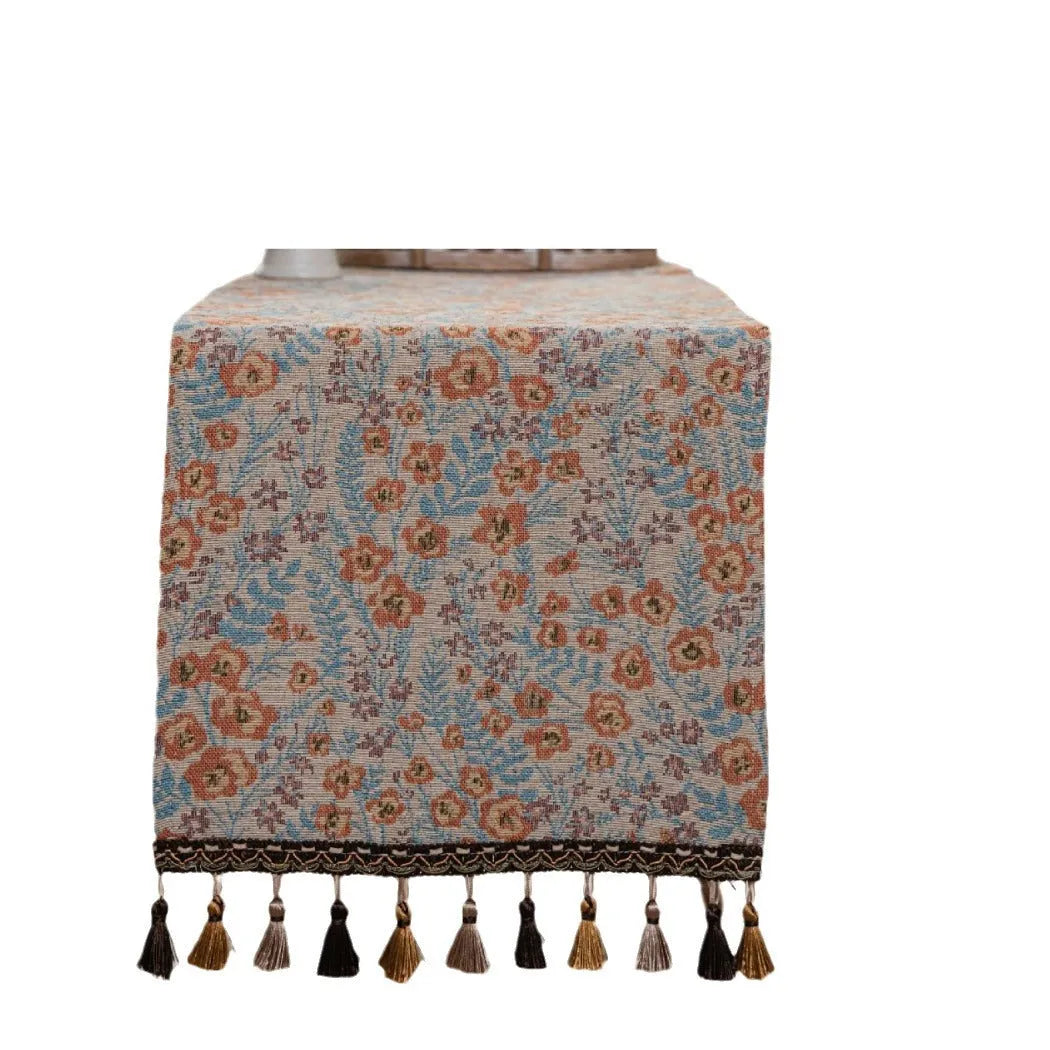 Beige polyester jacquard table runner with long tassels and a floral leaf pattern laid on a table.