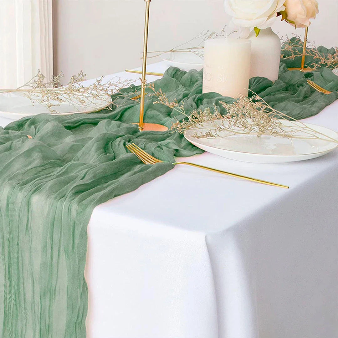 A green boho-style cheesecloth table runner with a delicate, wrinkled texture that adds a touch of elegant charm to any table setting.