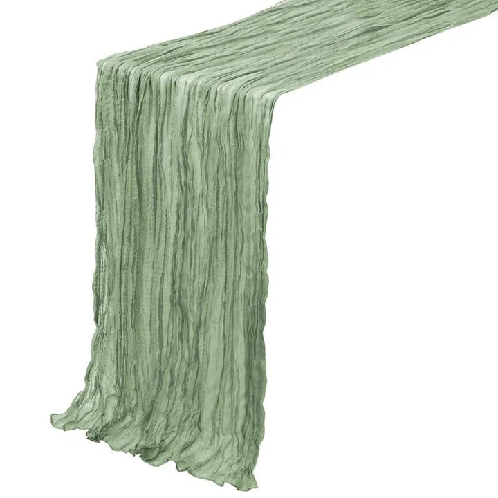 A green boho-style cheesecloth table runner with a delicate, wrinkled texture that adds a touch of elegant charm to any table setting.