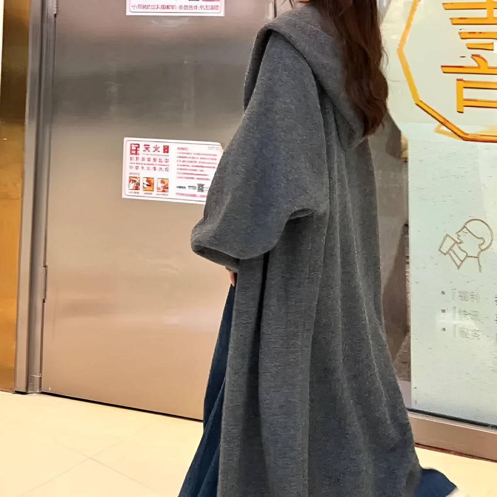 A woman wearing a long, beige, hooded knitted cardigan with a loose fit and below-the-knee length.