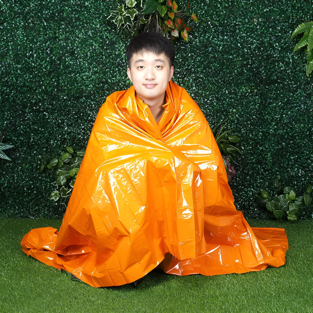Orange emergency survival blanket unfolded showing reflective inner surface, with folded sleeping bag and tent beside it in nylon bag