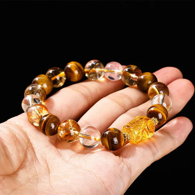 A close-up of a beaded bracelet with yellow banded Tiger's Eye and translucent citrine 10mm beads and a metallic Pi Xiu charm.
