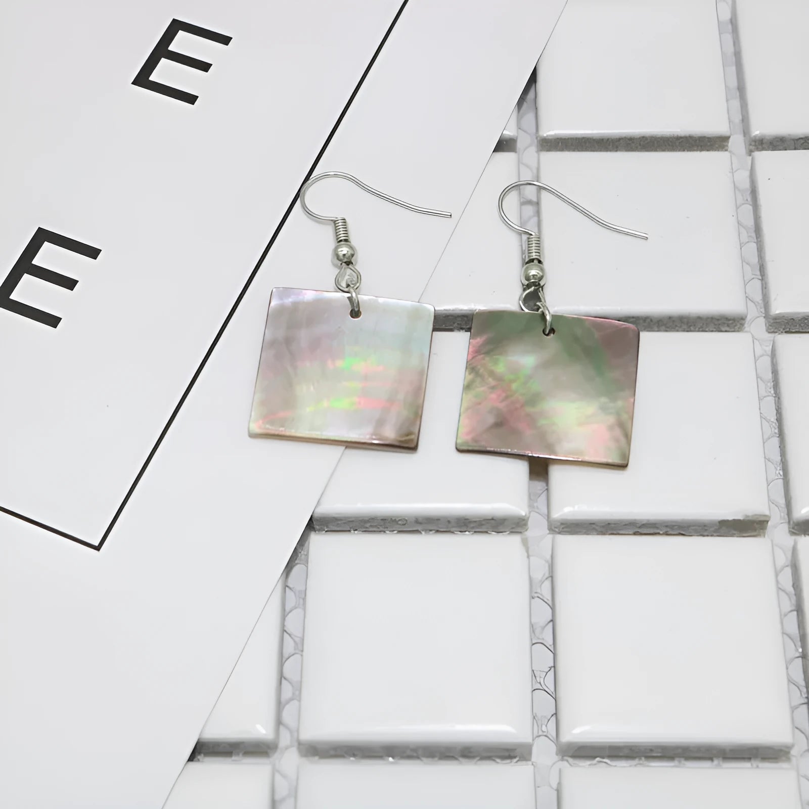 A pair of geometric shell earrings in a square panther design next to a rectangular shell design on a neutral background.