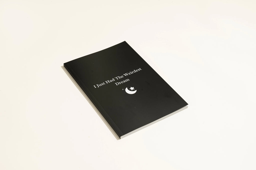 Black A5 hardcover dream journal with gold lettering, shown open on a lined page with a silk ribbon bookmark.