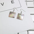 A pair of geometric shell earrings in a square panther design next to a rectangular shell design on a neutral background.