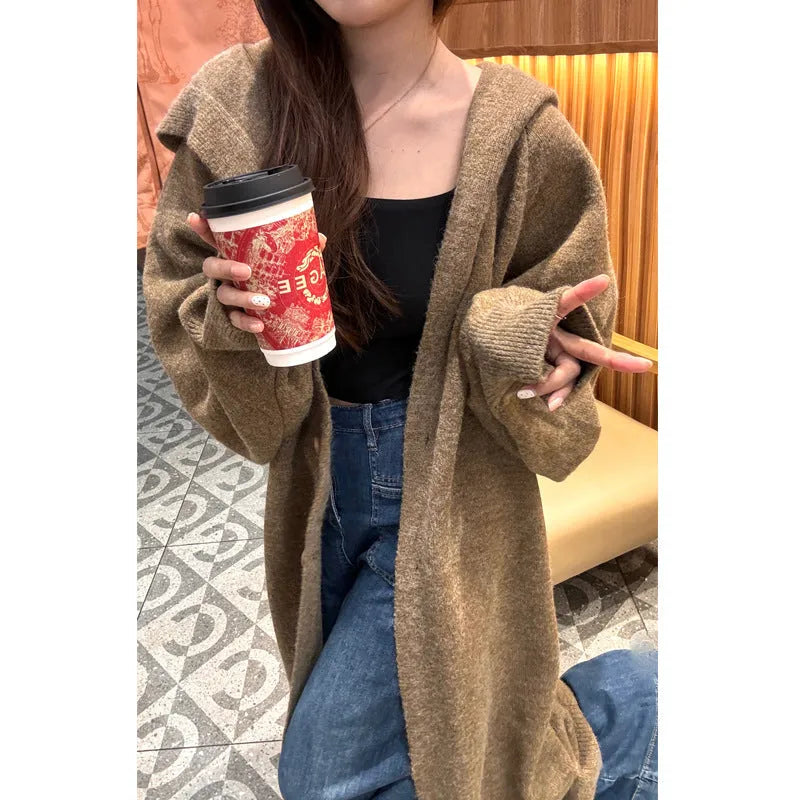 A woman wearing a long, beige, hooded knitted cardigan with a loose fit and below-the-knee length.