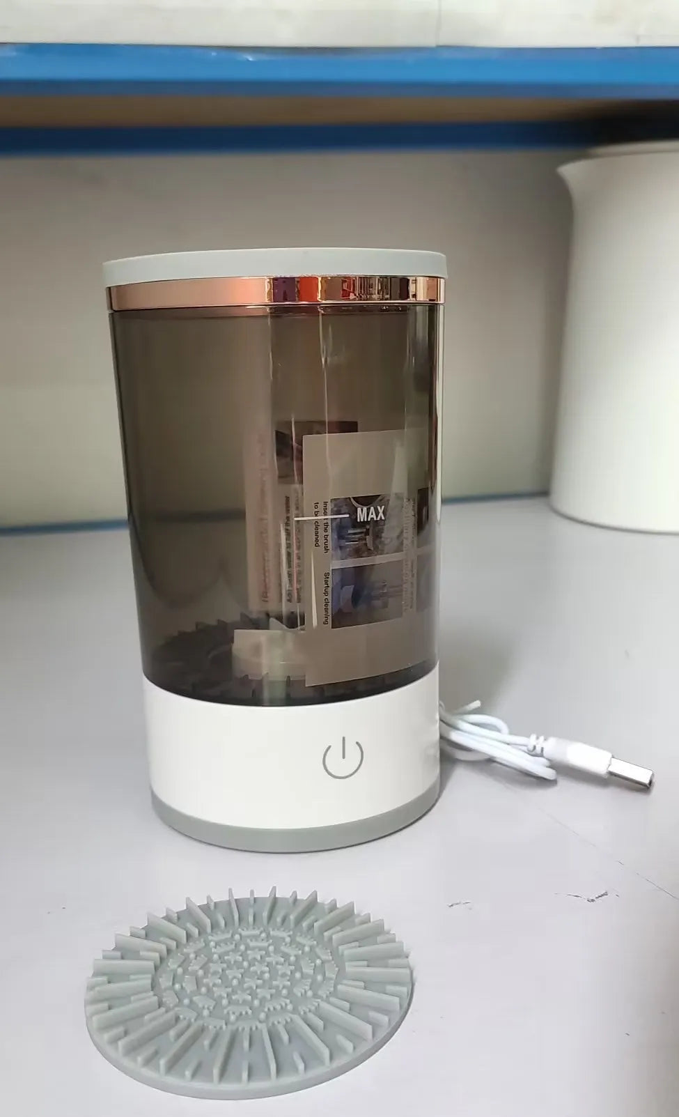 Grey and white electric makeup brush cleaner with USB charging cable on a wooden surface