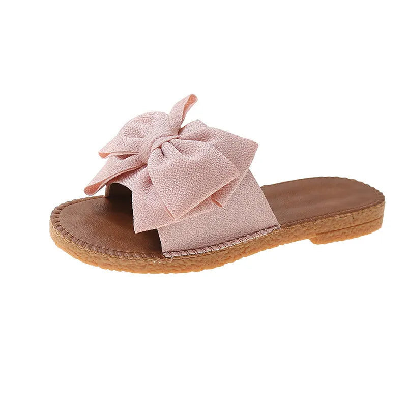 Women's beige suede slip-on sandals with a bow detail and a flat rubber sole.