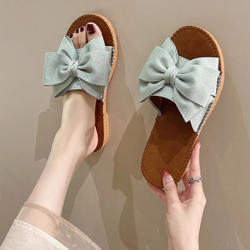 Women's beige suede slip-on sandals with a bow detail and a flat rubber sole.